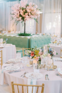 An elegant wedding reception table set with a light pink tablecloth, floral pattern plates, and a centerpiece featuring pastel roses, wildflowers, and glowing candles in glass cylinders. In the soft-focus background, more tables and a large flowering cherry blossom tree display sit within a ballroom garden setting.