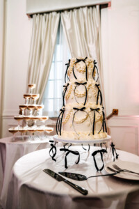 Three-tiered white wedding cake decorated with intricate cream piping and multiple thin black velvet bows. It sits on a round table next to a champagne glass tower and serving utensils in an elegant room with white curtains.
