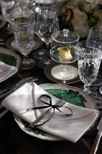 An elegant dinner place setting featuring a white napkin tied with a thin black bow, resting on a patterned plate with a green center and crystal glassware. In the background, a small butter dish is covered by a glass cloche amidst additional crystal wine and water glasses.
