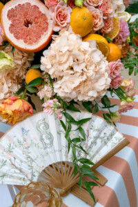 A paper hand fan with a floral design and the date July 13, 2024, rests on a peach and white striped tablecloth below a lush arrangement of grapefruit halves, oranges, and pink and peach roses.